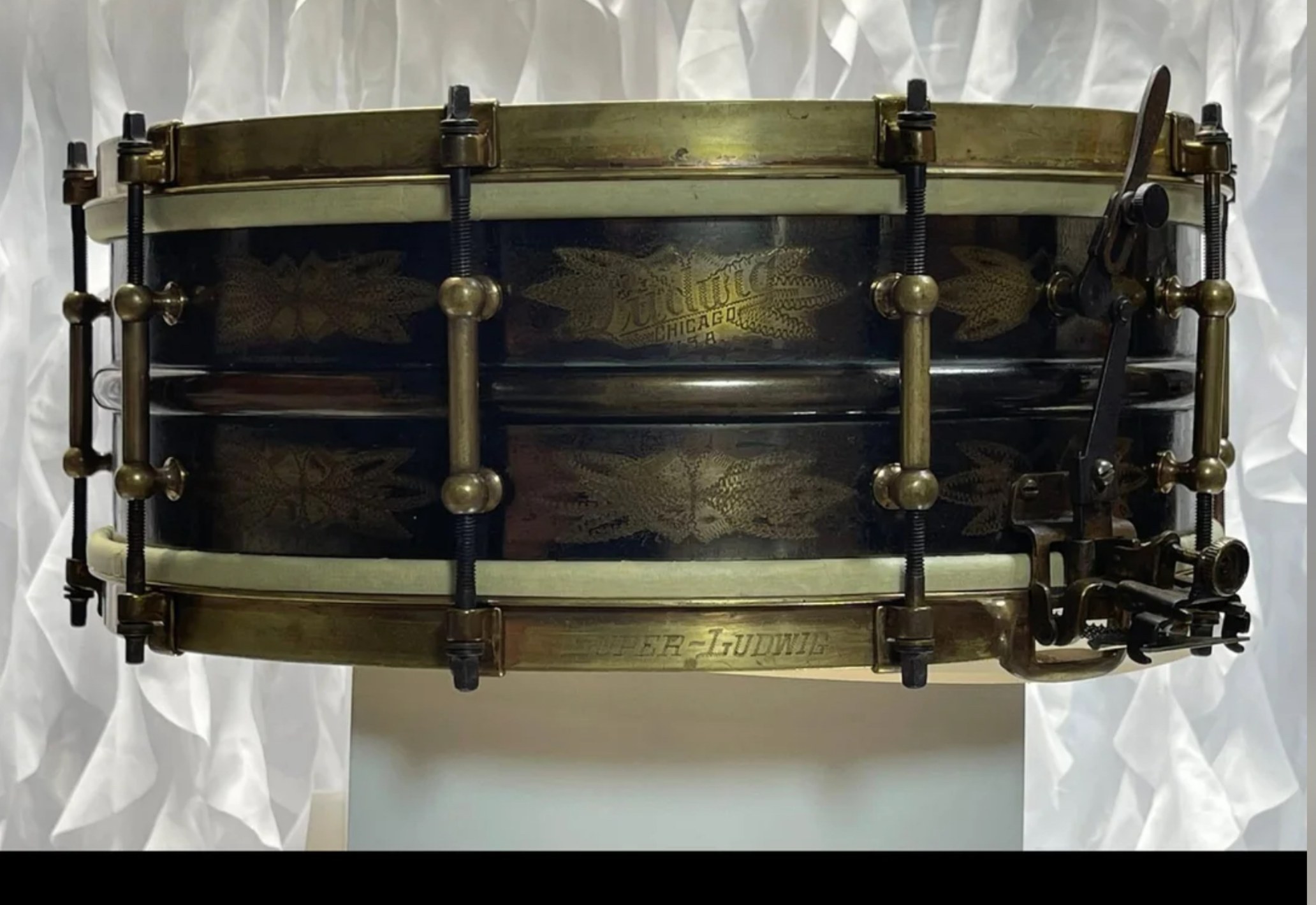 Restored vintage Super-Ludwig snare drum with engraved black shell, gold floral engraving, and aged brass hoops